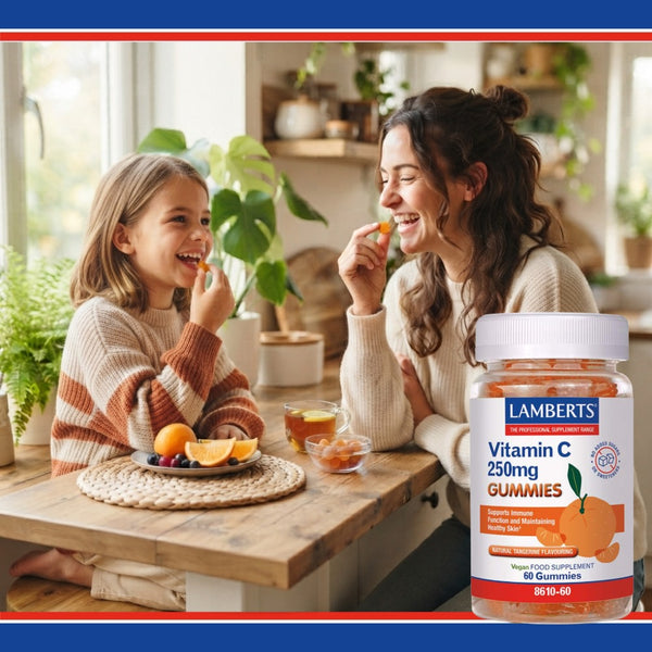 A woman and a child sitting at a wooden table in a bright kitchen, both eating vitamin C gummies. In the foreground, a large bottle of Lamberts Vitamin C 250mg Gummies is shown against a warm kitchen background with red and blue borders.