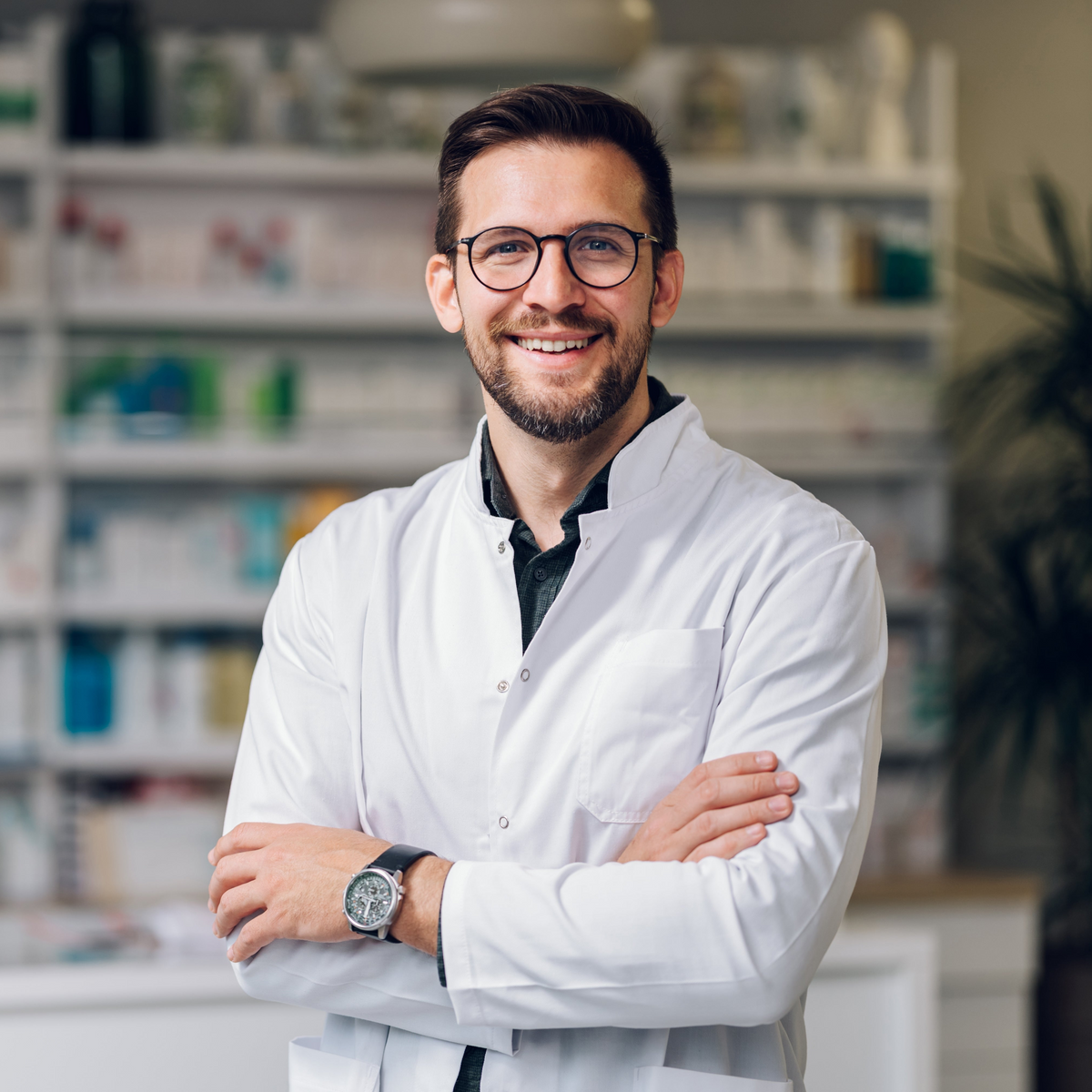 A smiling man wearing a white lab coat over a dark shirt, standing with arms crossed in front of a blurred pharmacy shelf background.