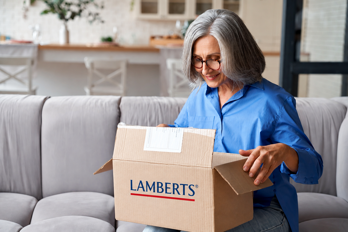 A woman with gray hair and glasses sits on a light gray couch, opening a cardboard box printed with the brand name LAMBERTS. The setting is a bright, modern living room.
