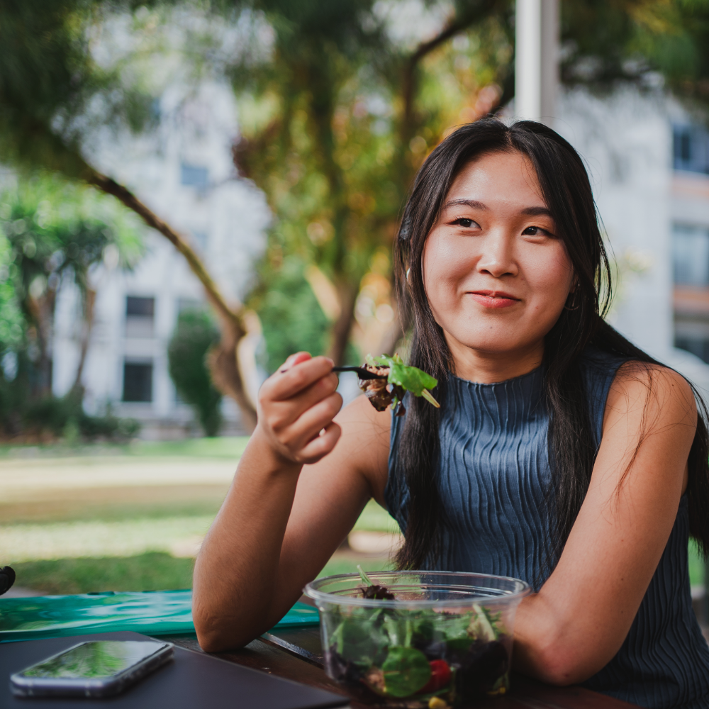 Woman eating a salad outdoors with trees and a house in the background