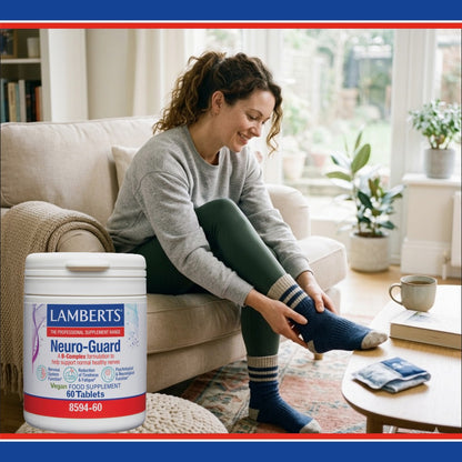 A woman sitting on a beige couch in a bright living room is putting on a blue striped sock while holding a large white tub of Lambert's Neuro-Guard supplement in the foreground.