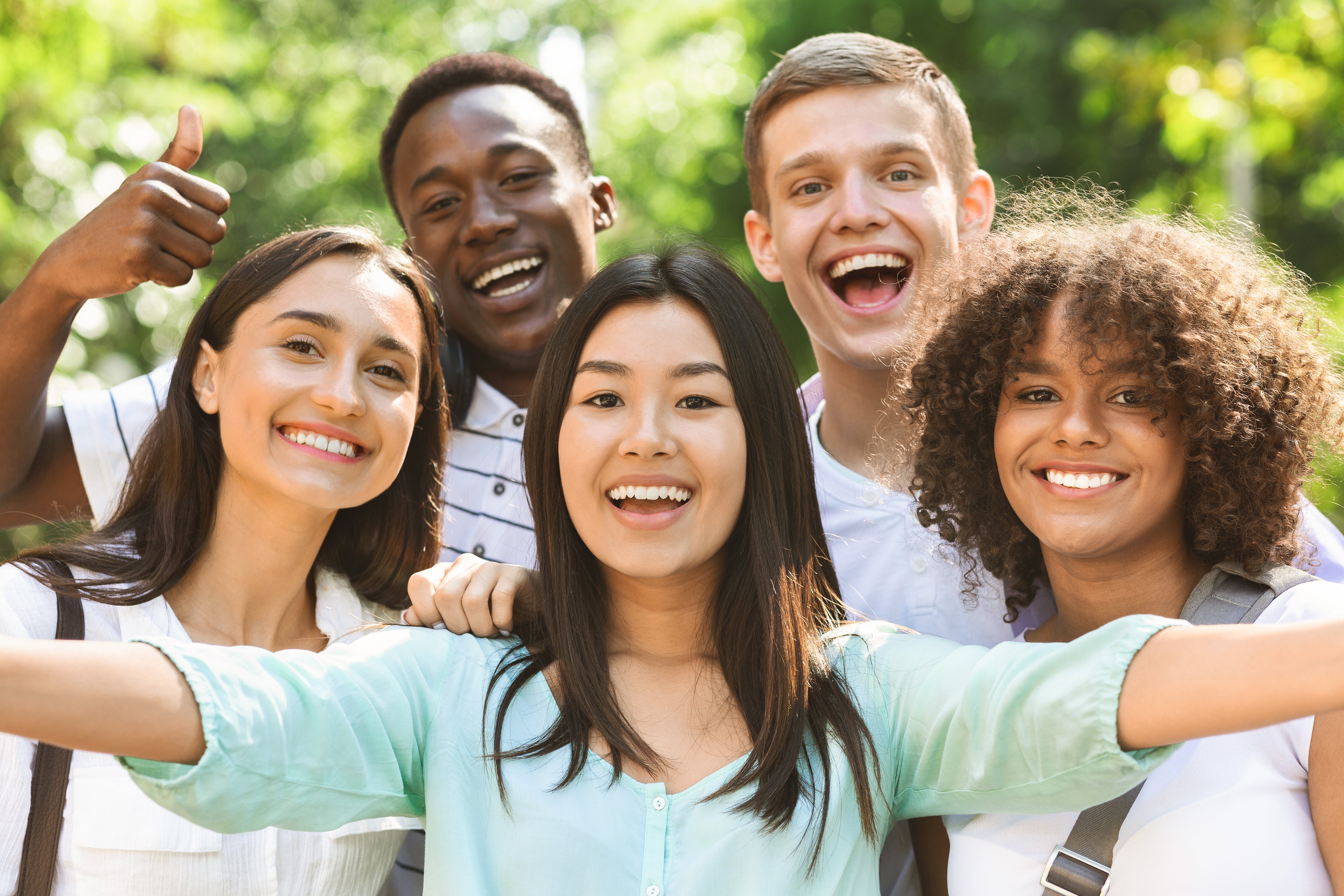 Group of five friends taking a selfie outdoors with greenery in the background